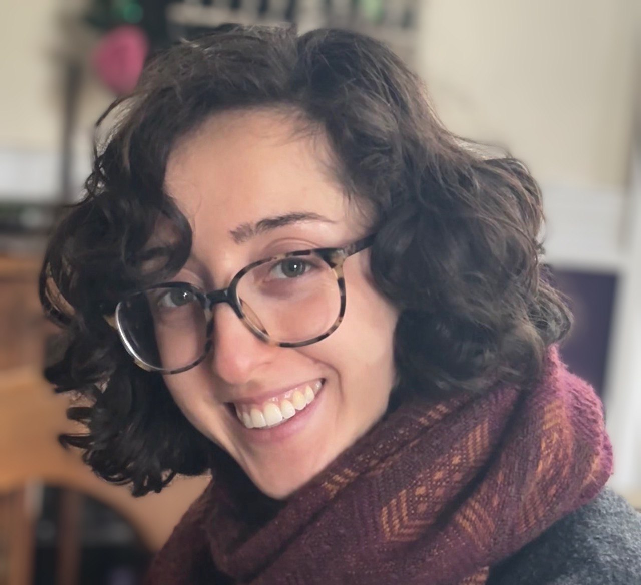 Amalya Megerman smiling in a red textured scarf and tortoise shell glasses, with a blurred background